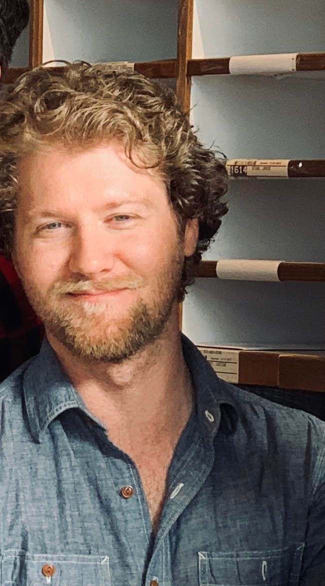 Smiling man in a blue denim shirt standing indoors in front of wooden shelves