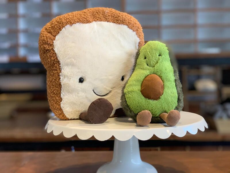 Plush toast and avocado toys on a white cake stand in a cozy café setting