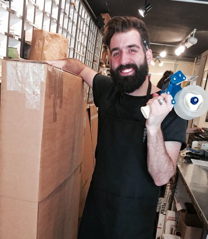 Smiling bearded worker in a black apron packing boxes beside a shelf in a warehouse setting