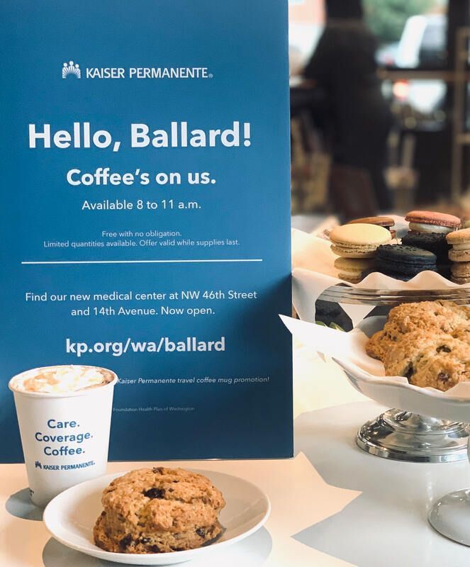 Coffee stand with pastries and a blue sign reading “Hello, Ballard! Coffee’s on us.”