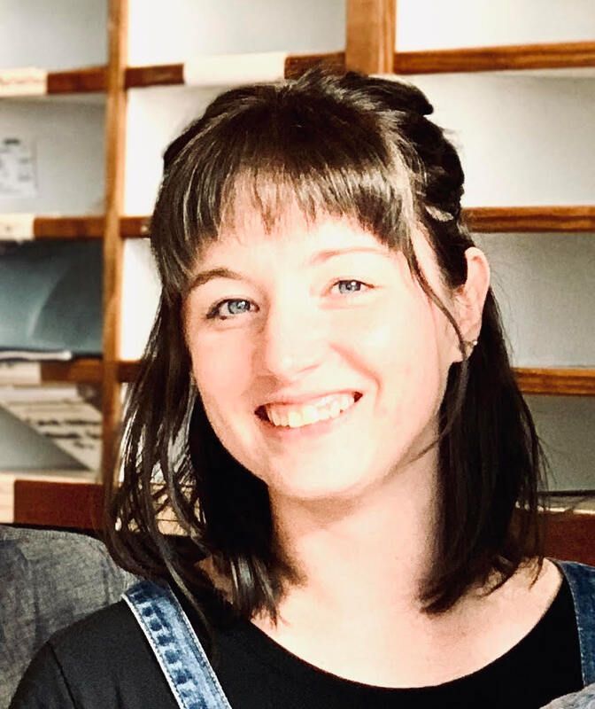 Smiling person with dark hair in a black top and denim overalls, sitting indoors against wooden shelves.