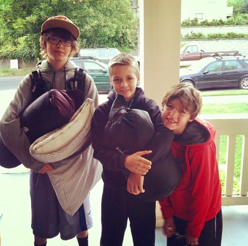 Three children on a porch holding blankets, smiling, with a street and parked car behind them.