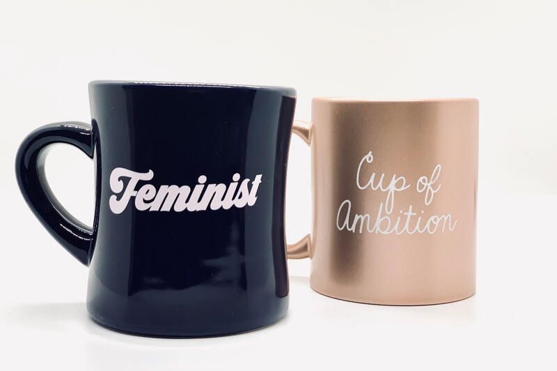 Two mugs with text: black mug “Feminist” and blush mug “Cup of Ambition”