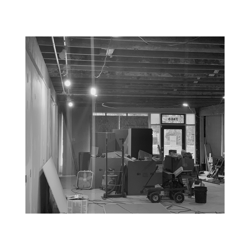 Black-and-white view of a cluttered unfinished room with exposed ceiling beams, equipment, and a doorway to outside