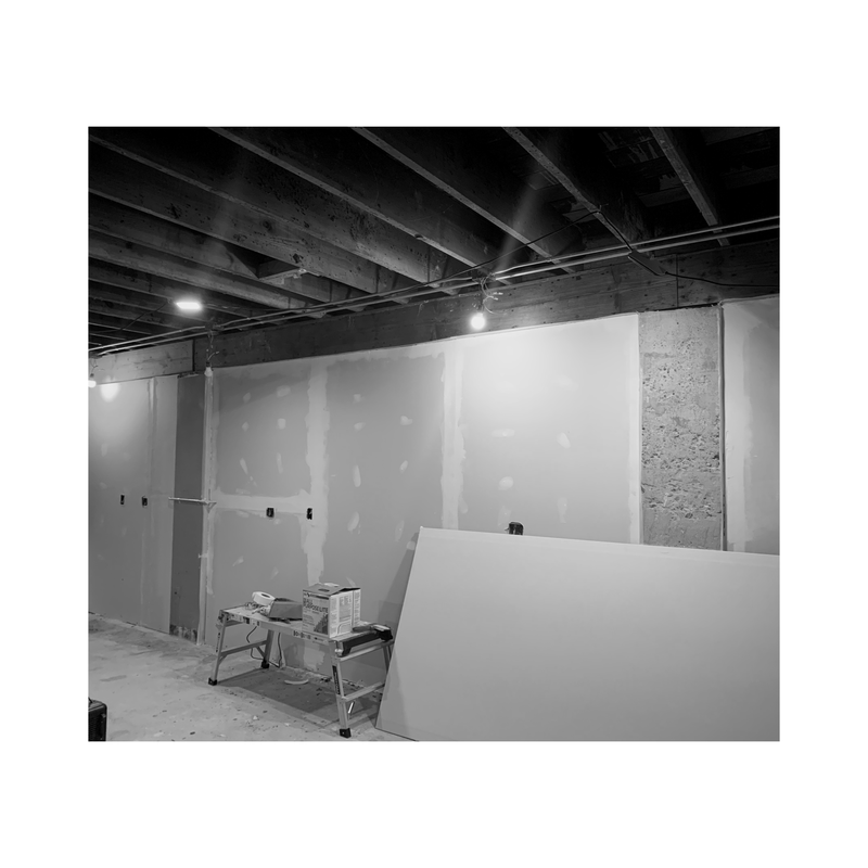 Black-and-white view of an unfinished room with drywall, exposed ceiling beams, and construction materials