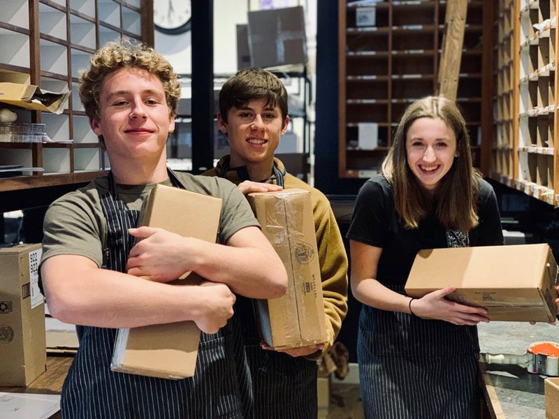 Three students stand in a workshop, smiling and holding cardboard boxes.