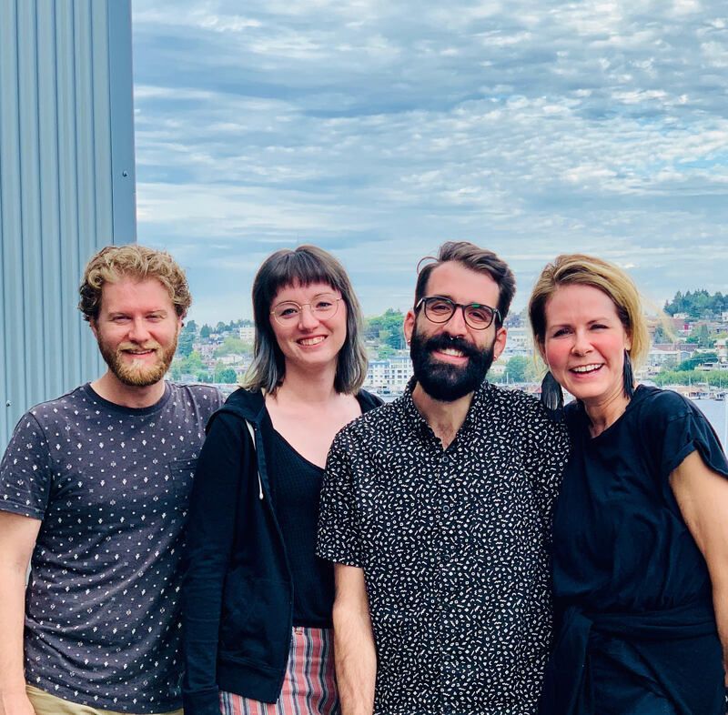 Four people smiling outdoors in front of a cloudy sky and water.