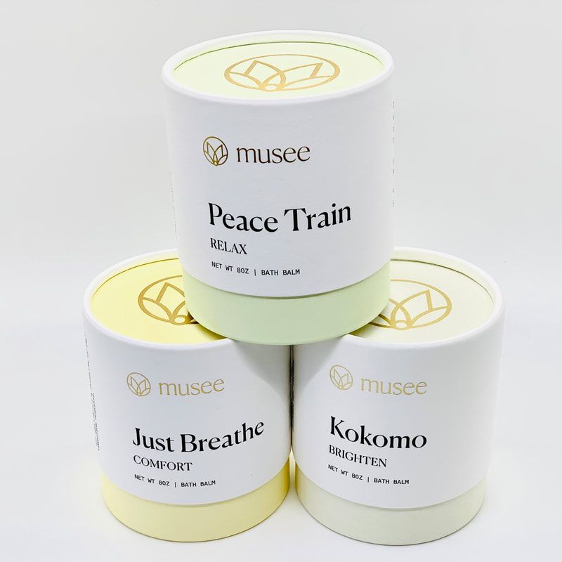 Three stacked Musee candle tins in white and pastel green/yellow, labeled Peace Train, Just Breathe, and Kokomo.