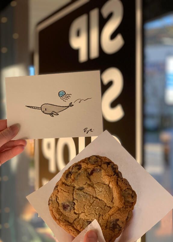 Hand holding a card and a cookie in front of a shop window sign.