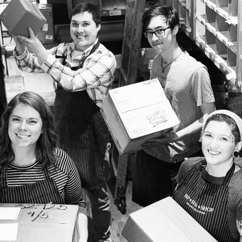Four workers in aprons packing boxes in a warehouse, smiling at the camera