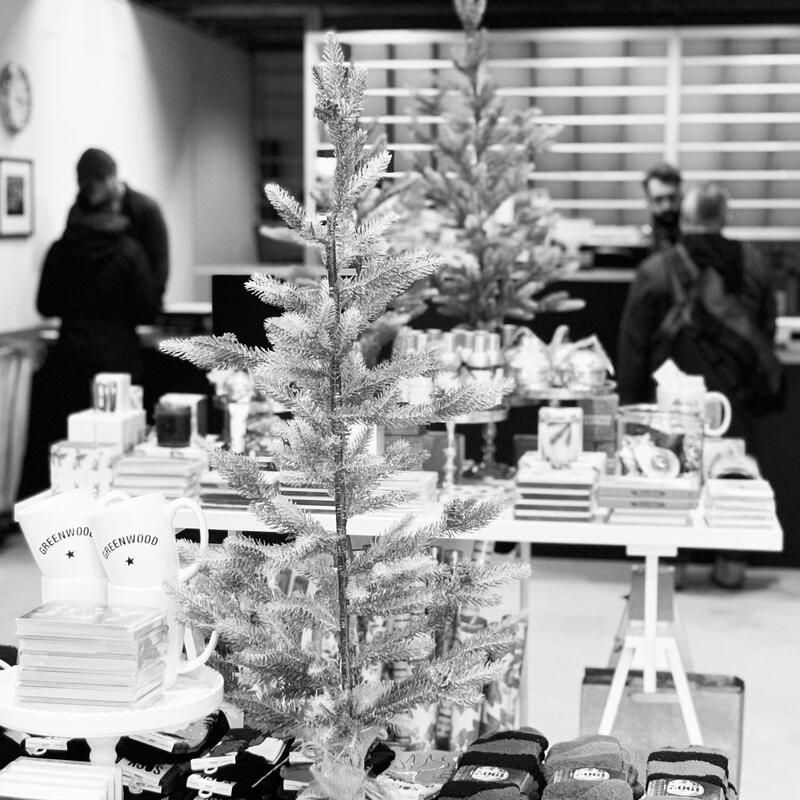 Small indoor holiday market with a decorated miniature Christmas tree in the foreground and people browsing tables.