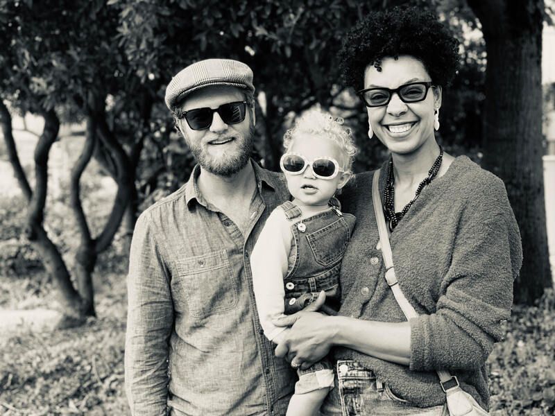 Three people smiling outdoors, with a child held between two adults.