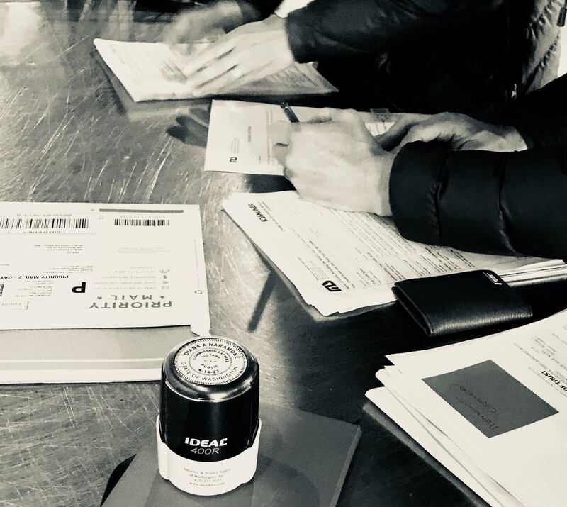 Hands sorting papers on a wooden table with a small jar in the foreground and stacks of documents nearby