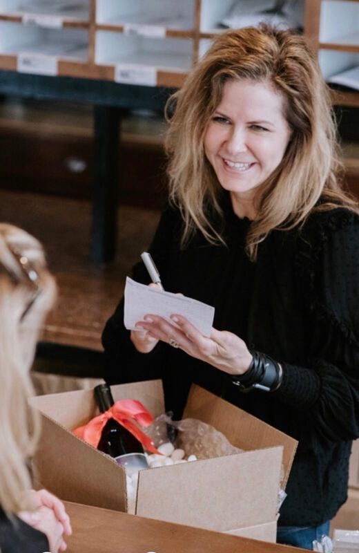 Woman smiling while holding a notepad beside an open box of items in a shop.