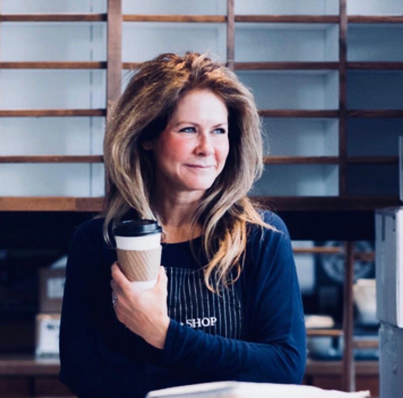 Woman holding a coffee cup in a shop, standing before wooden shelves and looking to the side.