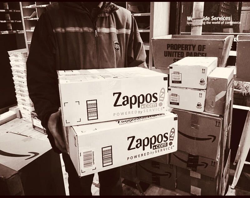 Person carrying two Zappos shipping boxes in a warehouse or loading area.