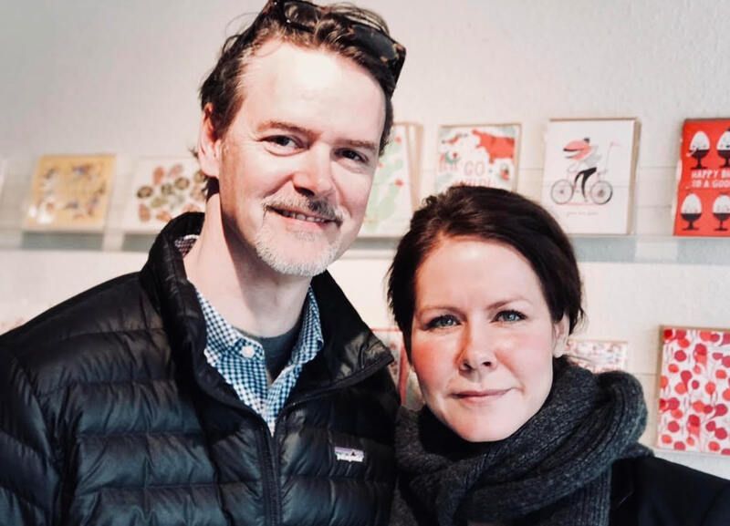 Two people smiling indoors, standing in front of colorful artwork displays.