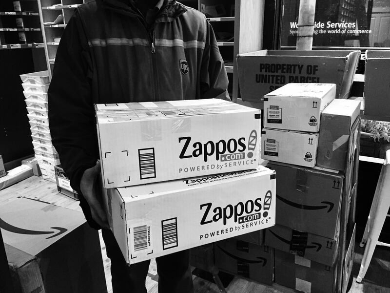 Person holding Zappos boxes in a warehouse, with stacked packages and a “Property of” sign.