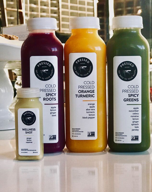 Four bottles of cold-pressed juice on a counter: one small beige bottle and three large red, orange, and green bottles.