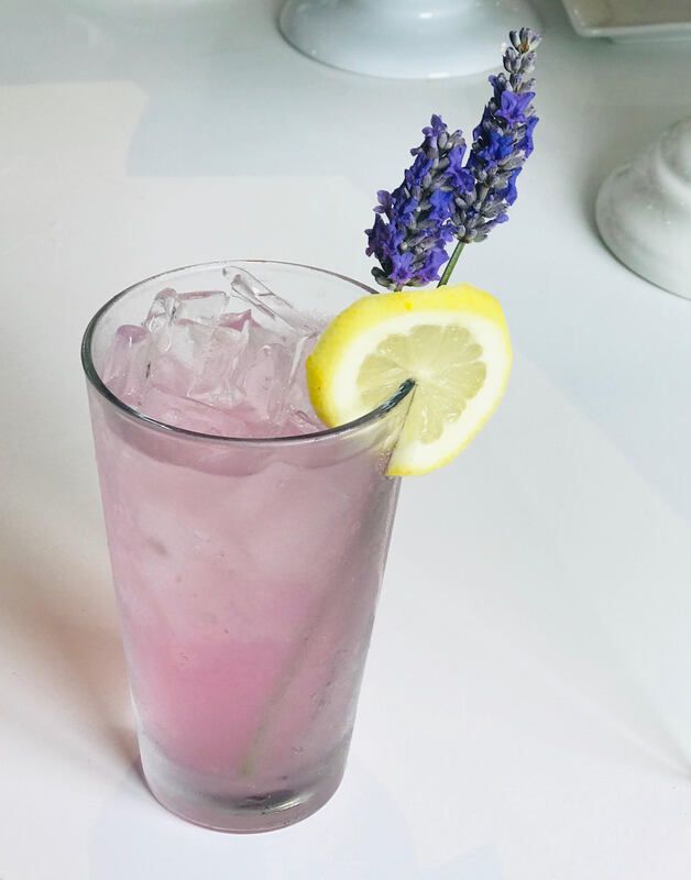 Pink iced drink with lemon slice and lavender garnish in a clear glass on a white table