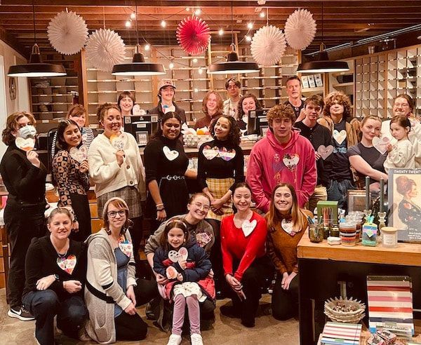 Large group posing in a warm kitchen, with a person in a bright pink hoodie front and center.