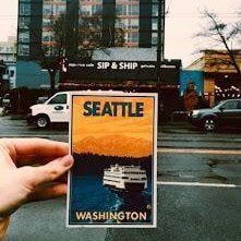 Hand holding a Seattle, Washington postcard on a city street with a ferry and “SIP & SHIP” sign in the background