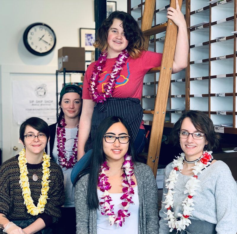 Five people indoors, smiling and wearing colorful leis, with one standing on a ladder in the center