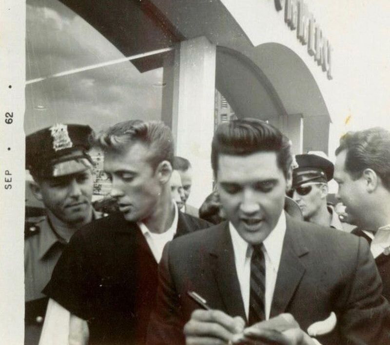 Elvis Presley signs autographs outside a building, surrounded by fans and police.