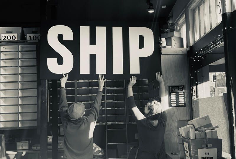 Two people in a warehouse raise a large “SHIP” sign overhead.