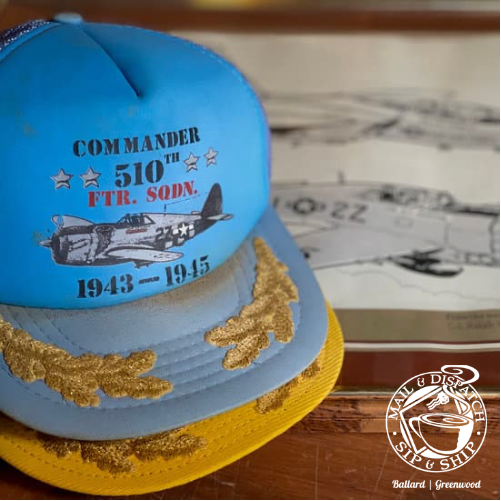 Blue and yellow commemorative cap with gold embroidery and navy aircraft graphic on a vintage map background