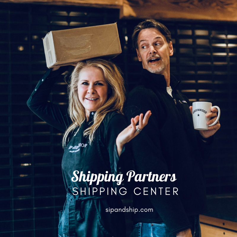 Two smiling people pose with a box and mug in front of a Shipping Partners sign