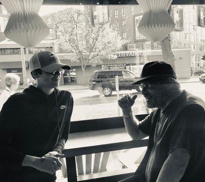 Two people talking in a cafe by a window, one in a cap and sunglasses, city street visible outside