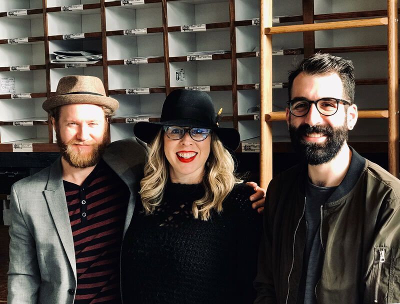 Three smiling people posing indoors in front of shelves, wearing hats and glasses.