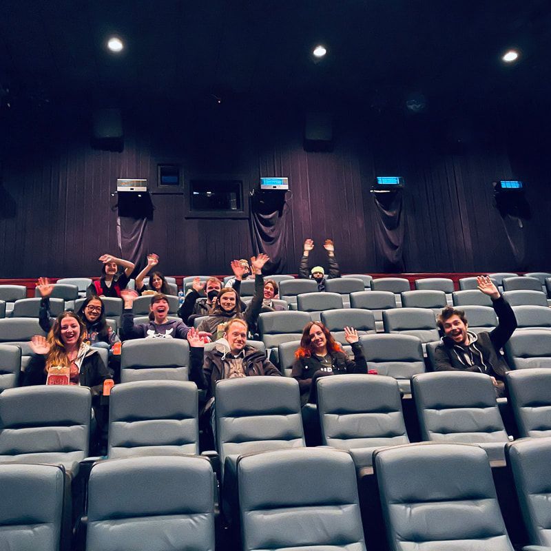 Small group seated in a dark movie theater, smiling and waving from several rows of gray seats