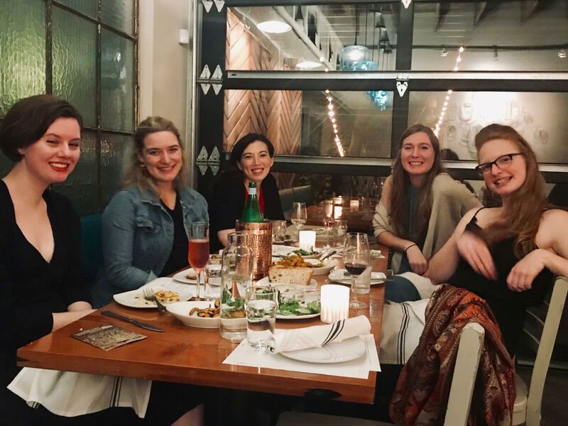Five friends smiling around a restaurant table with food, drinks, and a city view through the windows
