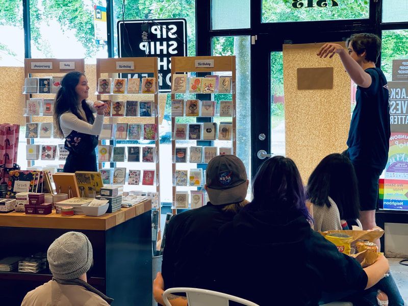 Two people presenting by a display wall in a shop or gallery, with seated attendees watching.