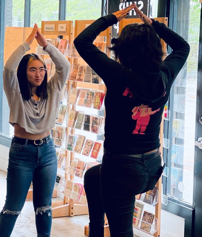 Two people pose with arms raised in a bookstore, standing near a display of greeting cards.