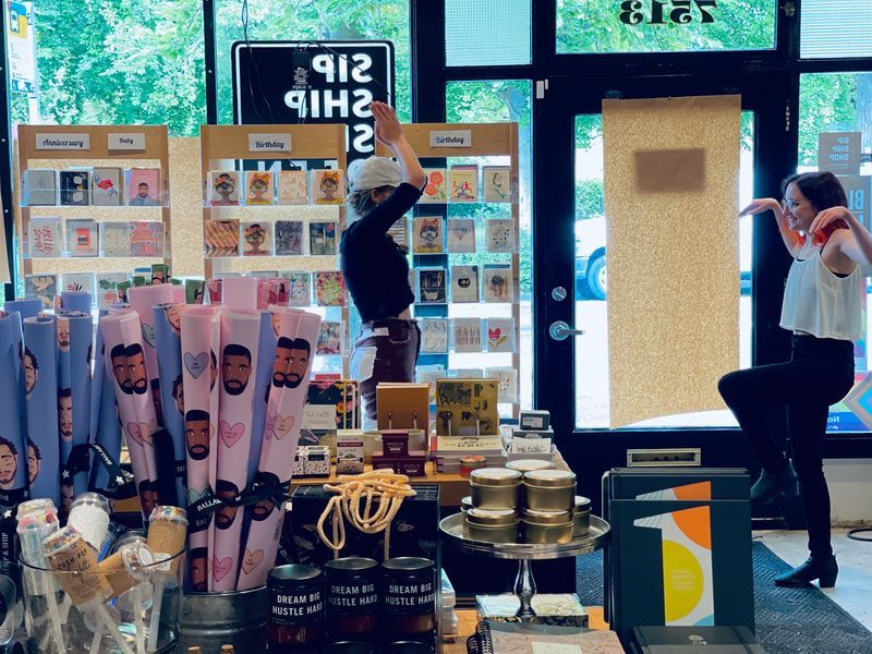 Shoppers in a brightly lit shop, browsing displays and phones near a counter of displayed goods.