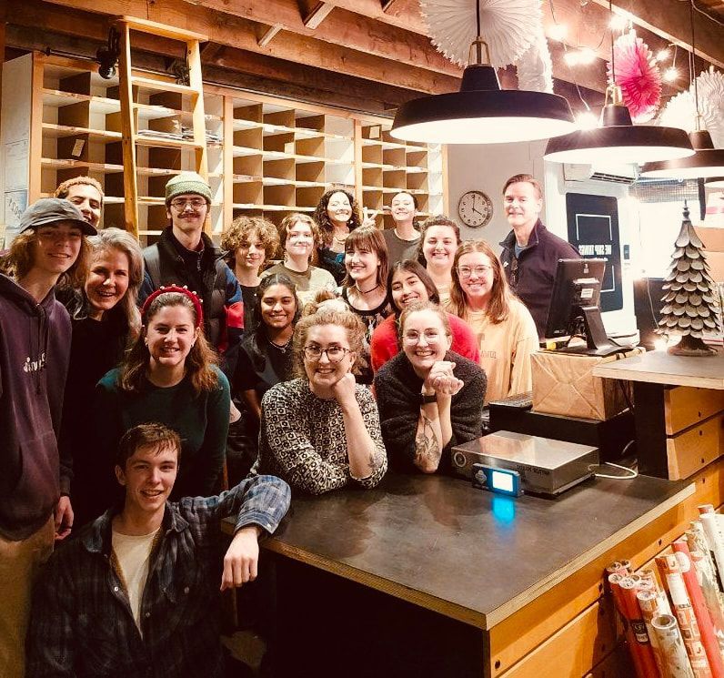 Group of smiling people gathered in a rustic café with shelves, wood beams, and warm hanging lights.