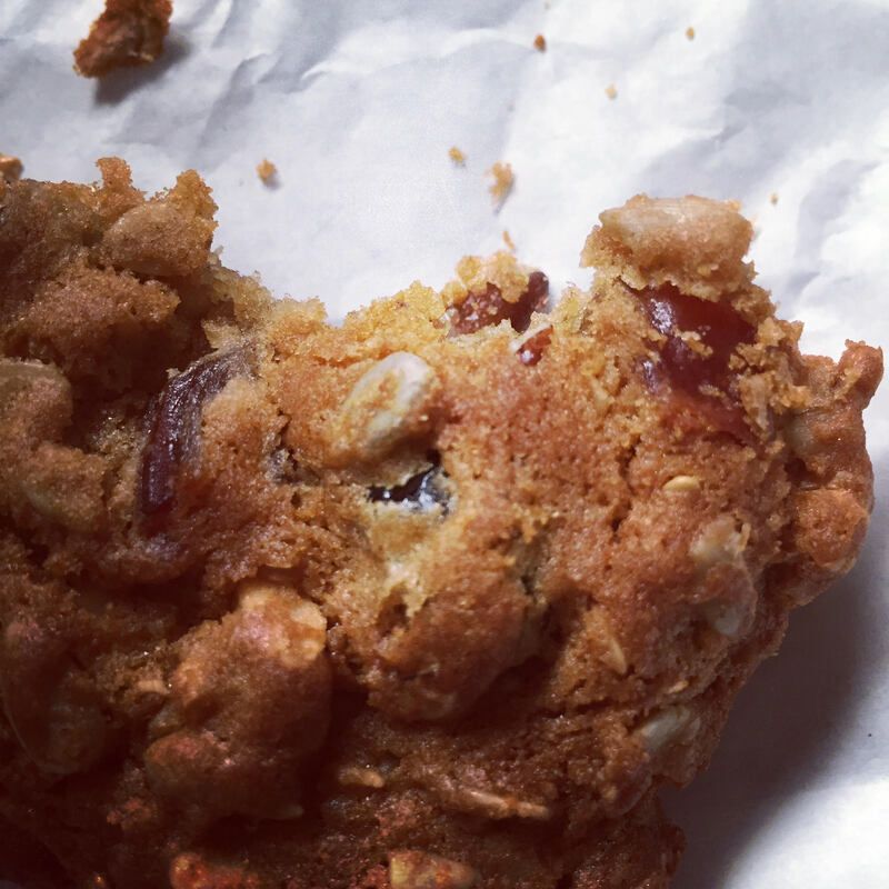 Close-up of a crumbly oatmeal cookie with visible raisins and nuts on white paper.