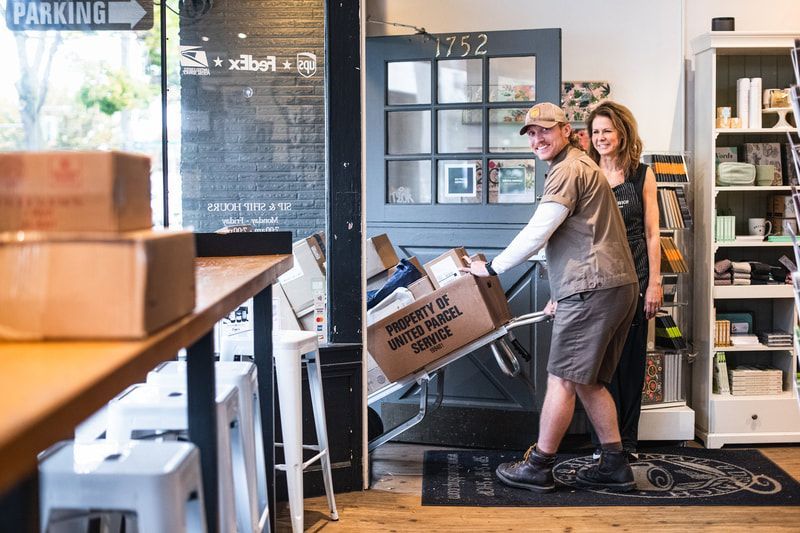 Two people moving a box truck inside a café with stacked shipping boxes nearby.