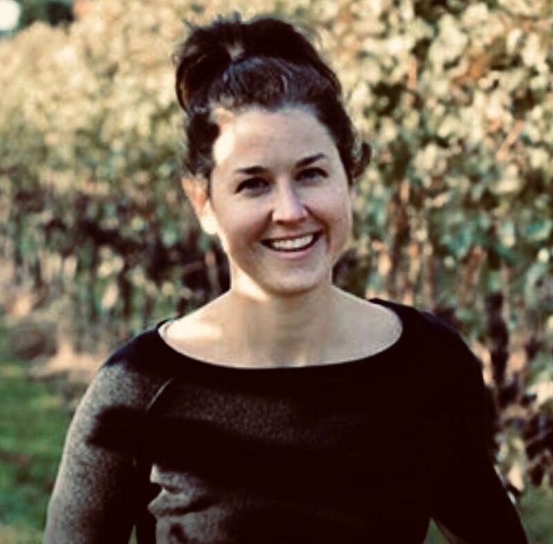 Smiling person with dark hair in a black top standing outdoors among trees