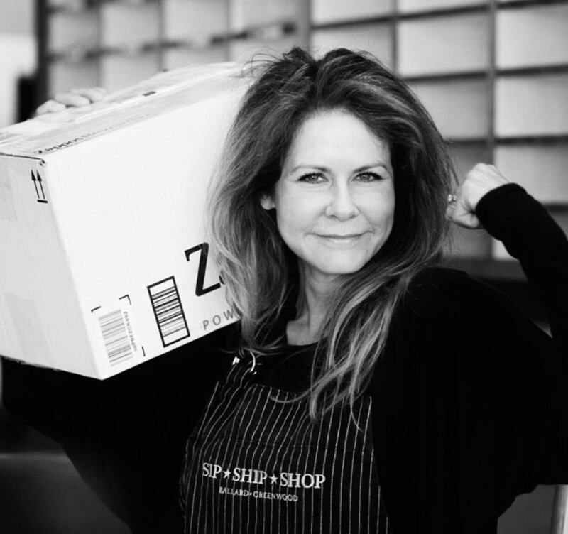Woman in an apron carrying a large box, standing indoors and smiling at the camera