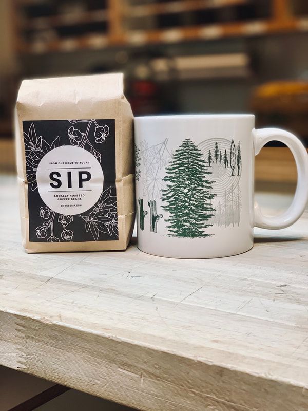 White mug with green trees beside a coffee bag labeled “SIP” on a wooden table.