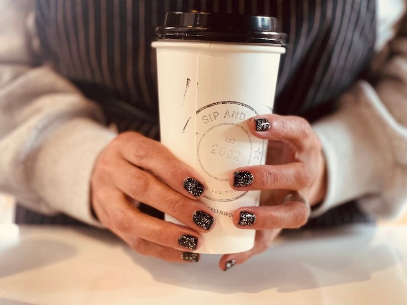 Hands with black glitter nails holding a white coffee cup with a black lid on a table.