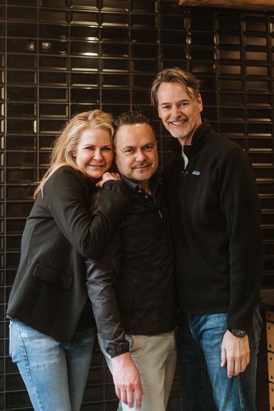 Three people smiling and posing together in front of a dark wooden wall.