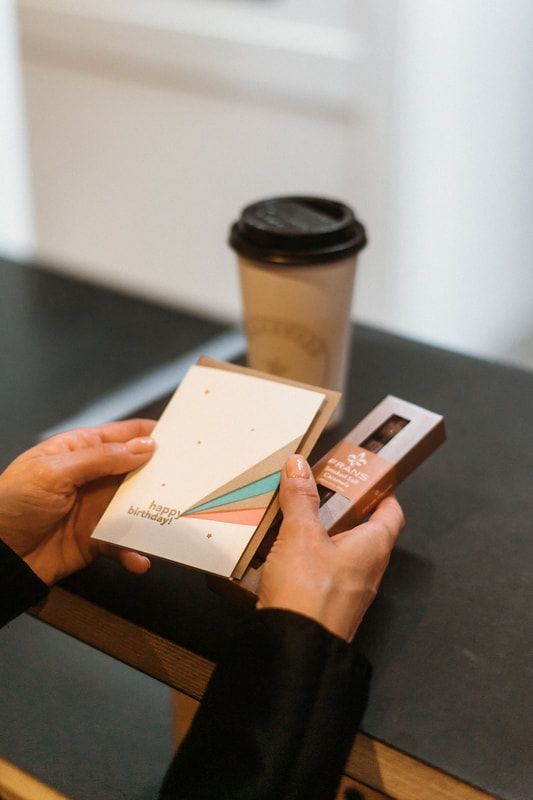 Hands holding a greeting card and chocolate bar beside a takeaway coffee cup on a table.