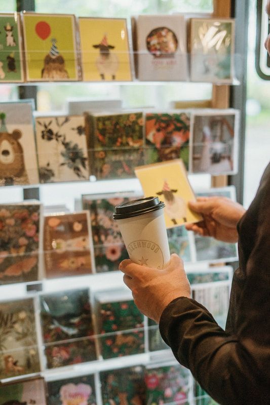 Person holding a coffee cup in front of a display of greeting cards and stationery.