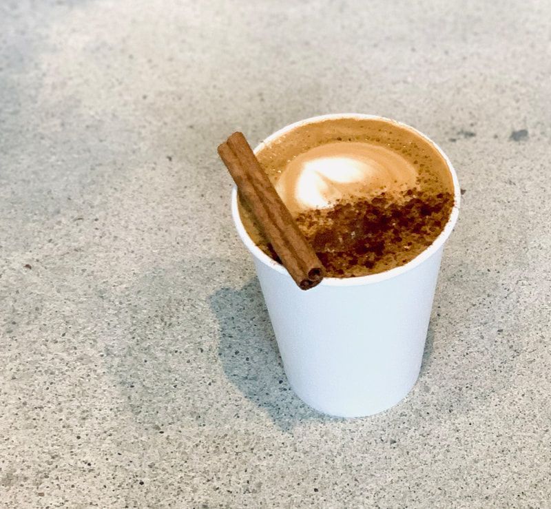 White paper cup of coffee topped with foam and two cinnamon sticks on a gray countertop