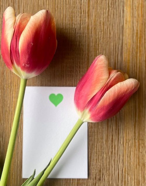 Two pink tulips beside a white card with a green heart on a wooden surface